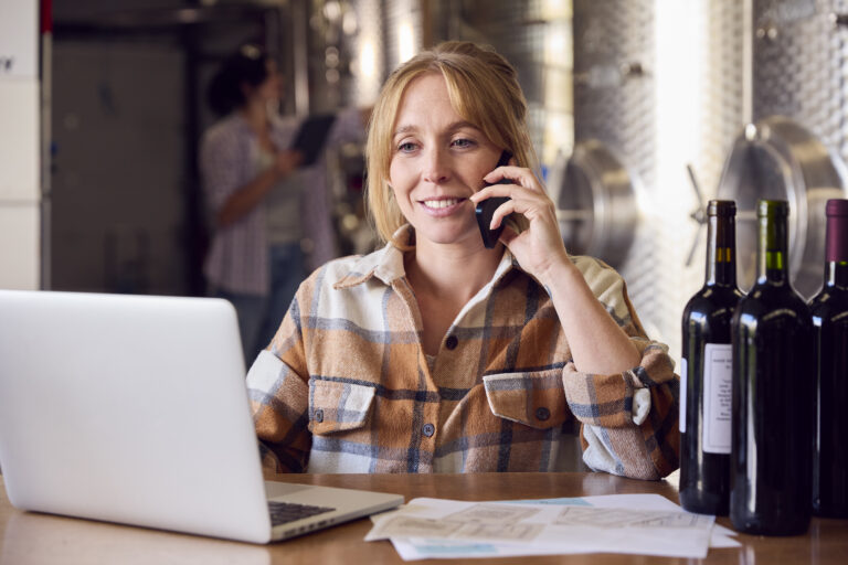 Woman On Mobile Phone With Laptop And Wine Bottles Inside Winery