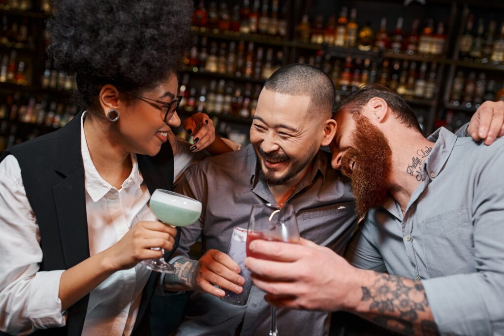 overjoyed multiethnic workmates with cocktails embracing and laughing in bar, leisure and fun