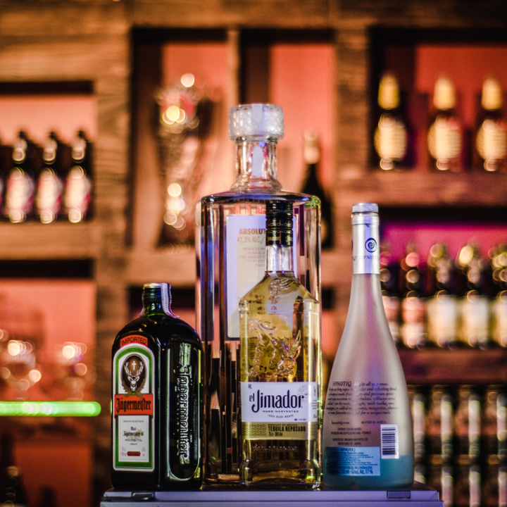 "A close-up of four liquor bottles displayed on a bar counter, including Jägermeister, El Jimador Tequila, Absolut Vodka, and a bottle of Hypnotiq. The background features a wooden bar shelf with illuminated bottles and glasses, creating a warm and inviting ambiance."