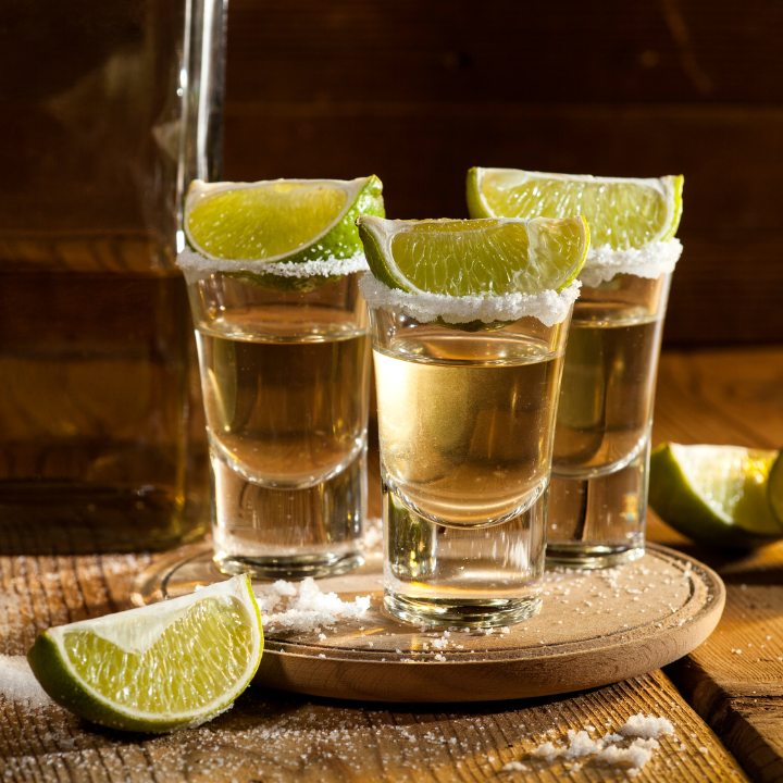 Three tequila shots with salted rims and fresh lime wedges, served on a wooden tray with scattered salt, set against a rustic background.