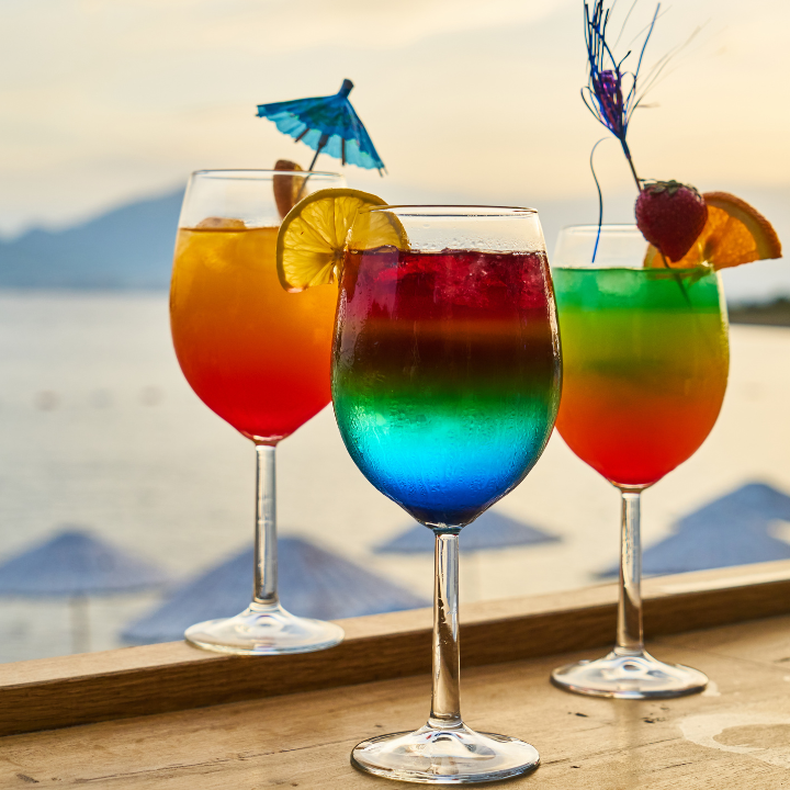 Three vibrant layered cocktails served in wine glasses, garnished with citrus slices, strawberries, and cocktail umbrellas, set against a scenic beach backdrop at sunset.