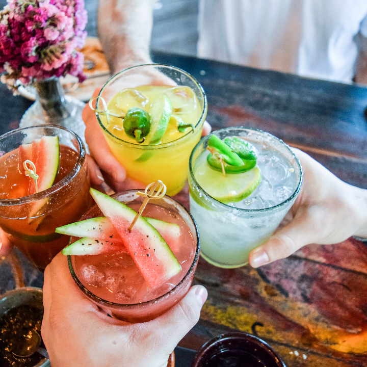 Four vibrant vodka-based cocktails garnished with fresh watermelon, lime, and jalapeños, being raised in a toast over a rustic wooden table.