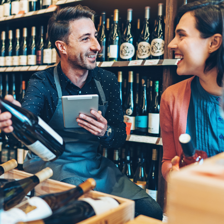 "A friendly wine store employee with an apron and tablet assisting a smiling female customer in selecting a bottle of wine, surrounded by shelves stocked with various wines."