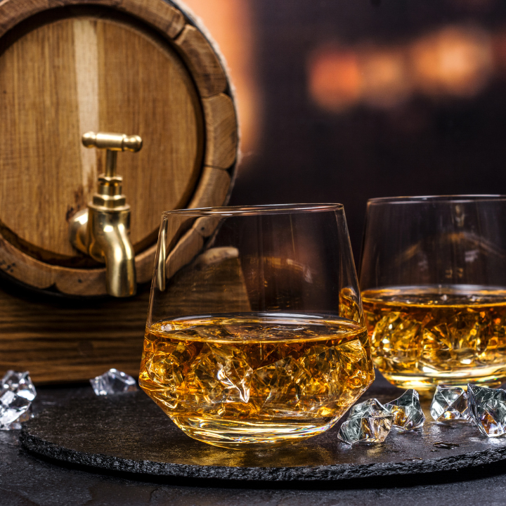 Two glasses of whiskey with ice cubes placed on a slate coaster, set against a wooden barrel with a brass tap, creating a warm and elegant ambiance.