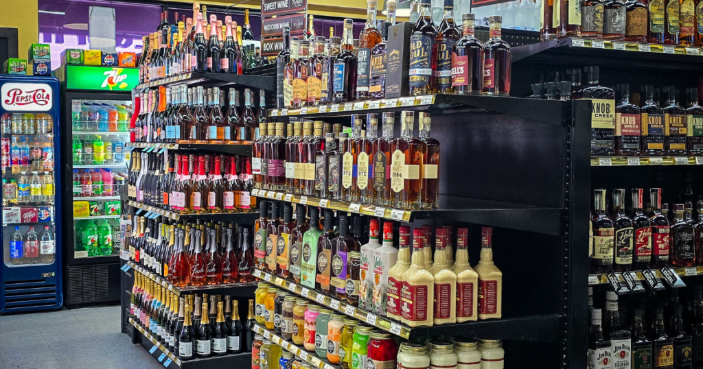 Welcoming liquor store aisles neatly stocked with premium wines, spirits, and liqueurs; chilled soft drinks like Pepsi in back.